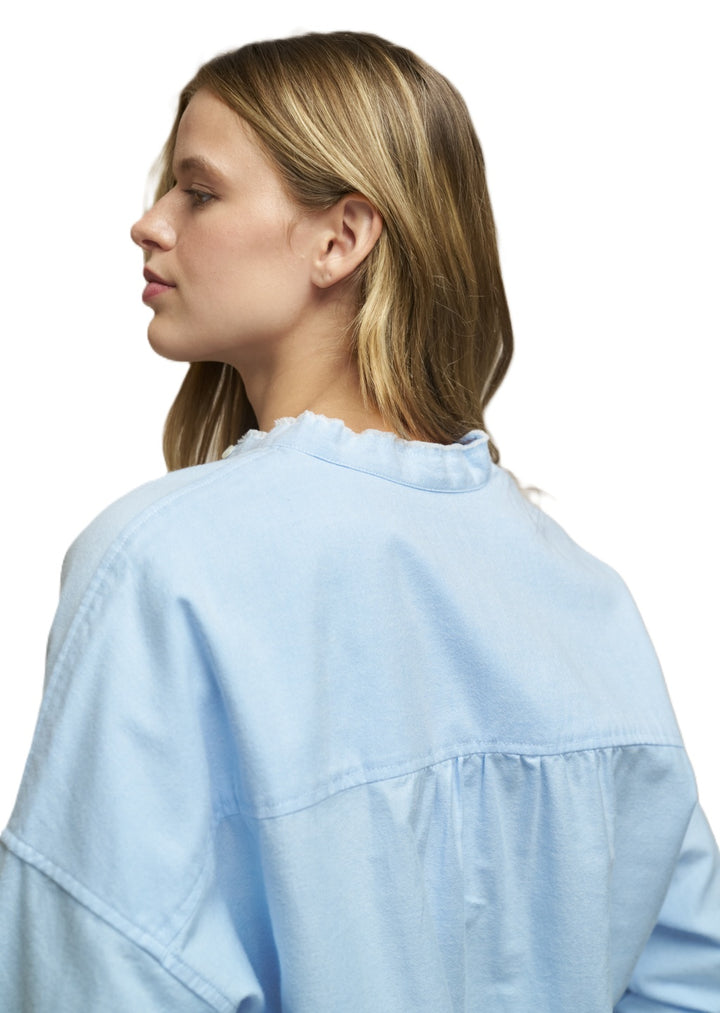 Bluse 0039 Italy Lucy RAW in hellblau. Super hübsche Bluse mit geradem Schnitt und überschnittenen Schultern. Die Bluse hat einen flachen Stehkragen mit Rüschendetails. Sie hat eine durchgehende Knopfleiste. Die Ärmel sind lang und haben eine Einknopf-Manschette. Die Rückenpartie ist verlängert und der Saum ist abgerundet. 