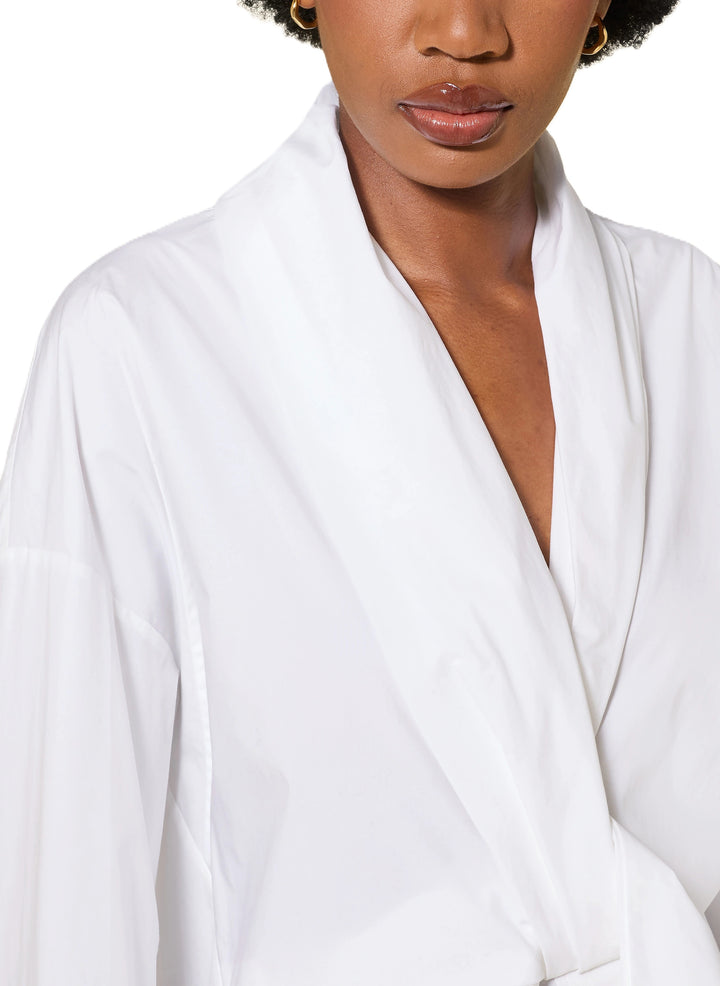 Bluse Dorothee Schumacher POPLIN POWER blouse in weiß. Diese Bluse ist ein richtiges It-Piece. Sie vereint modische Wickeloptik mit femininem Schnitt perfekt. Die Bluse hat einen Schalkragen und lange Ärmel. An den Manschetten befinden sich schöne Knöpfe. Die Ärmel sind weit und die Schultern überschnitten. Auf der Rückseite ist die Bluse eingeschlagen und der Saum mit einem elastischen Band versehen.