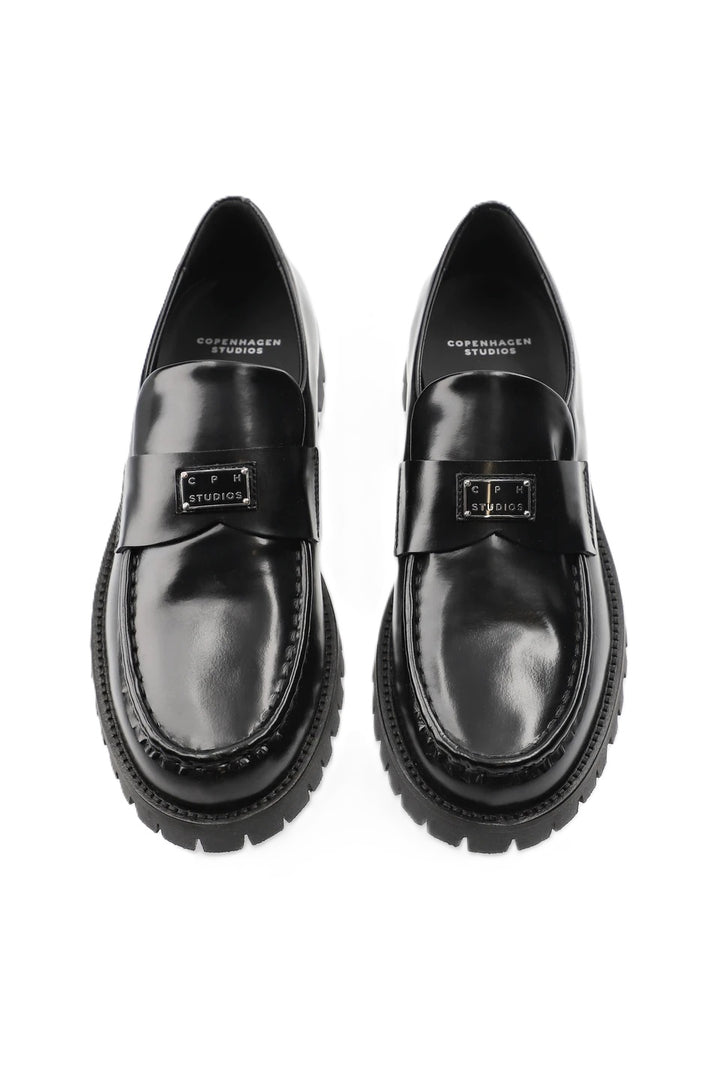 Schuh Copenhagen Studios CPH848 in schwarz. Dieser Loafer ist schmal geschnitten und hat eine glatte, klare Form. Der V-förmige Steg, das glänzende, gebürstete Finish und das Logopatch aus Metall auf der Zunge machen den besonderen Look aus. Das Leder ist sehr weich und der Schuh sehr angenehm zu tragen. 