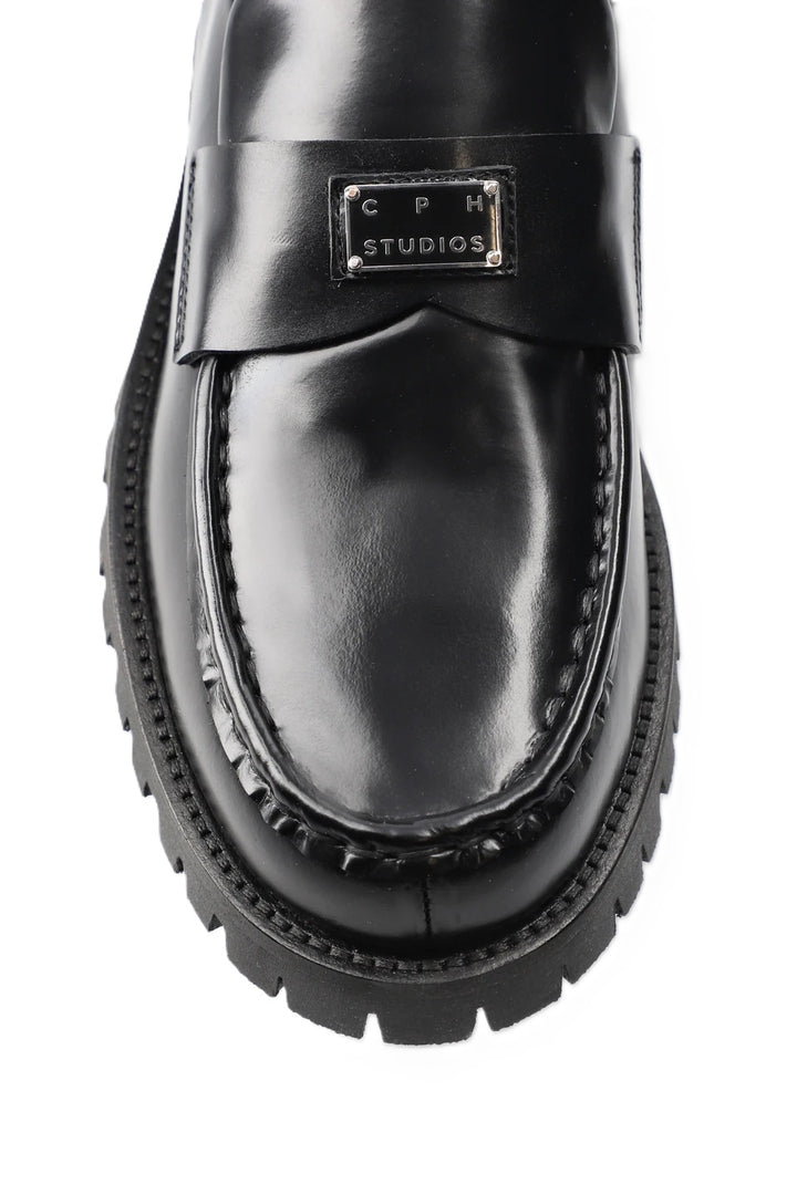 Schuh Copenhagen Studios CPH848 in schwarz. Dieser Loafer ist schmal geschnitten und hat eine glatte, klare Form. Der V-förmige Steg, das glänzende, gebürstete Finish und das Logopatch aus Metall auf der Zunge machen den besonderen Look aus. Das Leder ist sehr weich und der Schuh sehr angenehm zu tragen. 