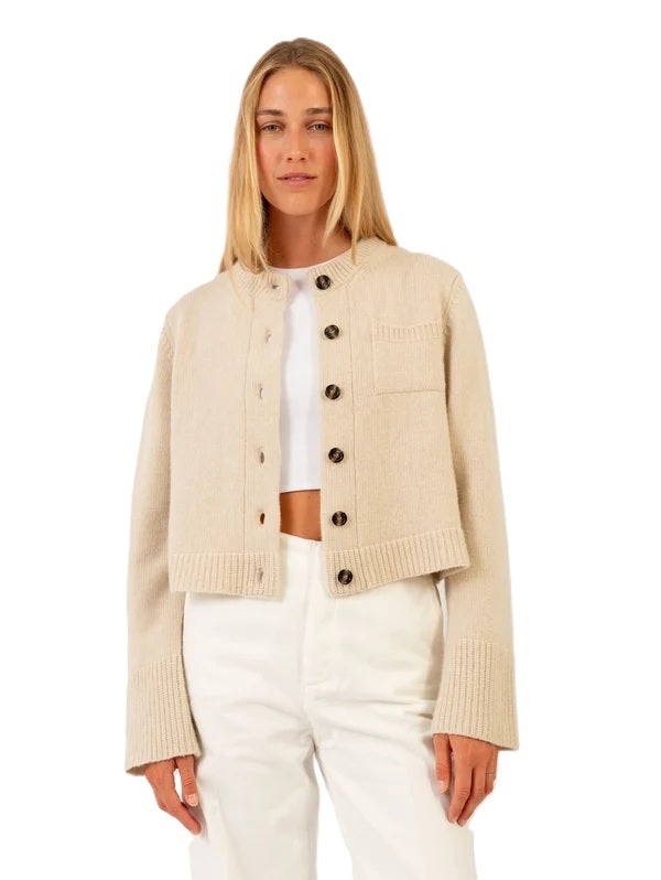 Strickjacke Anna V Berg Lilly Jacket buttermilk. Hübscher Cardigan in beige. Der Cardigan hat einen Stehkragen und farblich abgesetzte Knöpfe. Auf der einen Seite befindet sich eine kleine Brusttasche. Die Säume sind mit Rippstrick abgesetzt. 